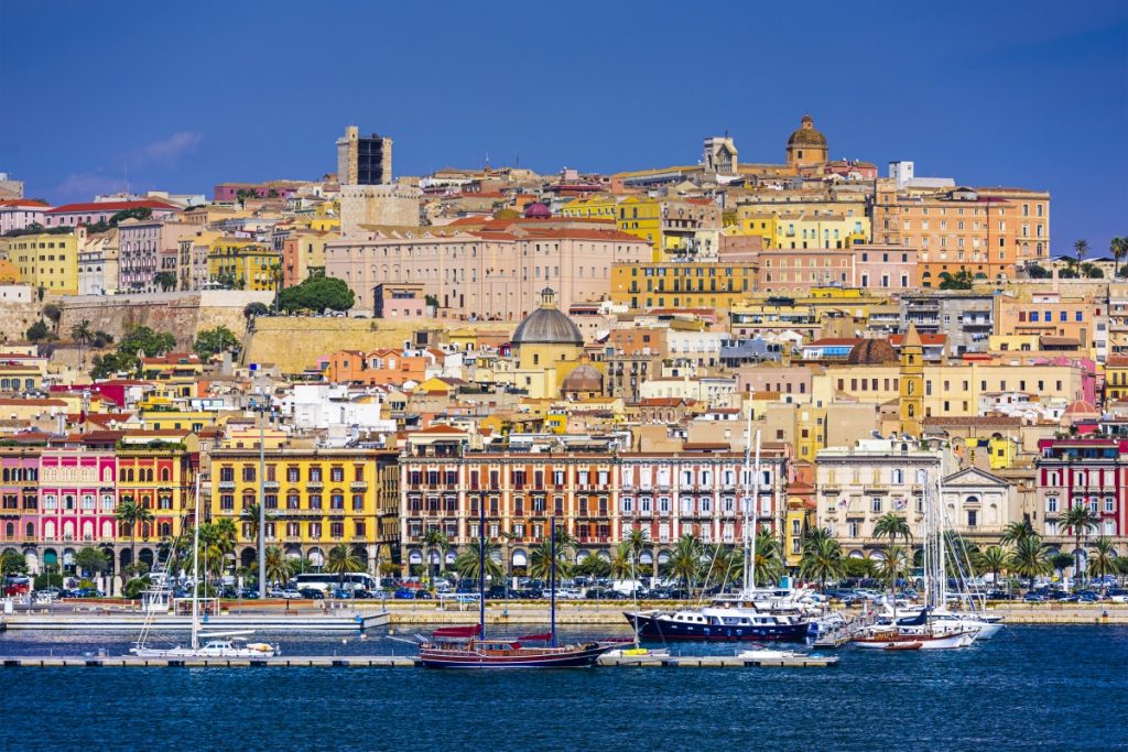 Cagliari skyline viewed from the sea, with colorful buildings rising above the harbor and historic landmarks visible on the hill.