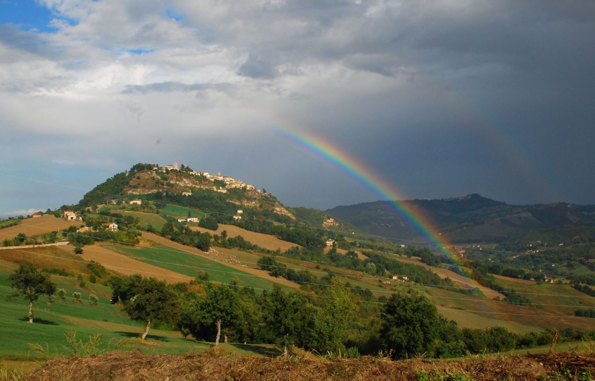 Monte San Martino village nestled on a hillside with a vibrant rainbow arching across the sky above the surrounding green landscape.