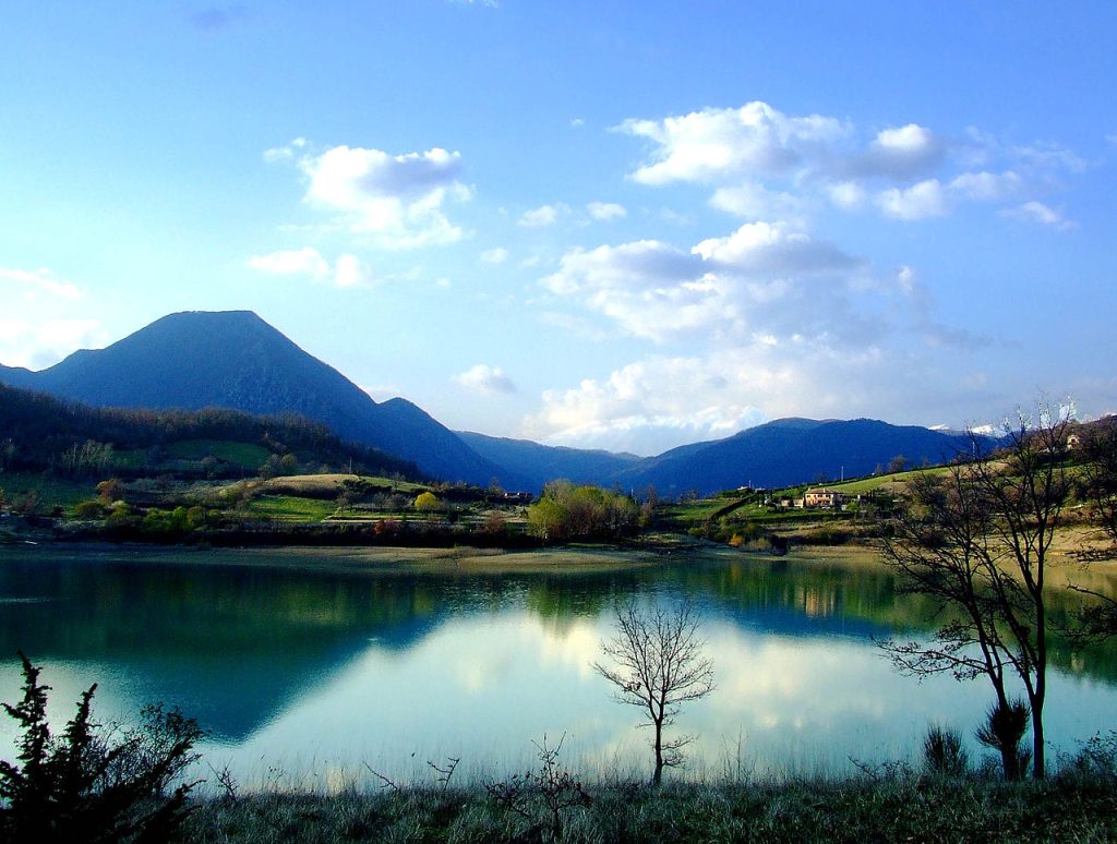 Panoramic view of Lake Castel San Vincenzo in Molise, Italy, with turquoise water, forested hills, and the Apennine mountains in the distance.
