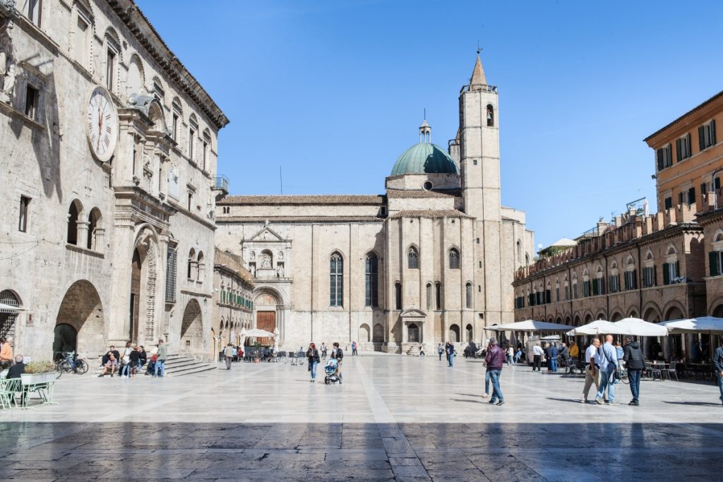 Piazza del Popolo in Ascoli Piceno, featuring the elegant travertine buildings, colonnades, and the clocktower under a bright sky.