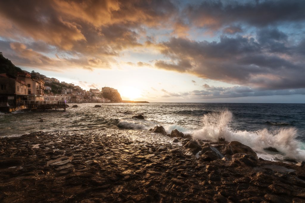 Sunset view of Scilla with waves crashing against the shore and the cliffside village glowing in warm light.
