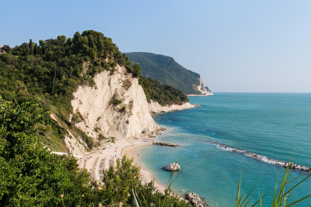 A scenic view of Sirolo perched on the cliffs overlooking the turquoise waters of the Riviera del Conero, captured for a Marche travel guide, with limestone rocks and lush greenery framing the coastline.