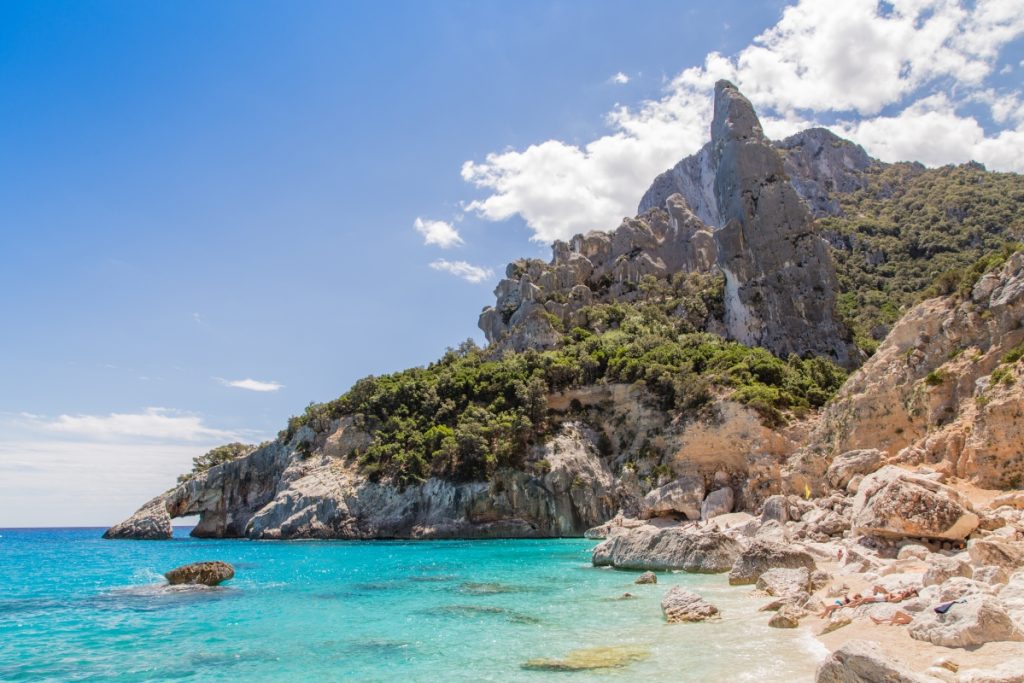 Spiaggia di Cala Goloritzé with turquoise water, white pebbles, and limestone cliffs, featuring the iconic rock pinnacle by the shore.