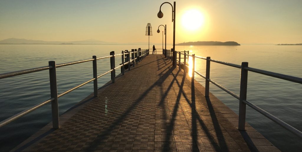 View of Passignano sul Trasimeno’s lakeside village with stone buildings along the waterfront and boats on Lake Trasimeno under a soft sunset.